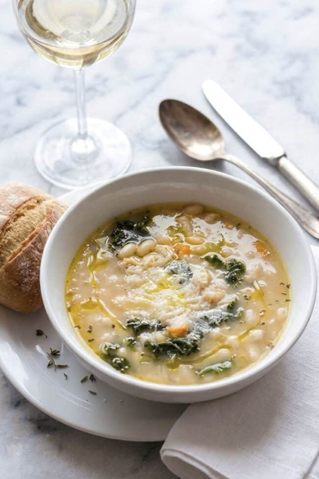A bowl of Tuscan white bean and kale soup garnished with olive oil and parmesan cheese, served with a piece of crusty bread and a glass of white wine.