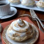 A small pumpkin tartlet topped with toasted swirls of meringue sits on a decorative plate with a fork and napkin nearby. In the background, more pumpkin tartlets and a cup of coffee are visible.