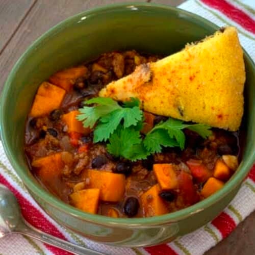 A green bowl of turkey chili with yams, black beans, sweet potatoes, and tomato pieces, garnished with fresh cilantro and a slice of cornbread. The bowl sits on a striped cloth napkin with a spoon beside it.