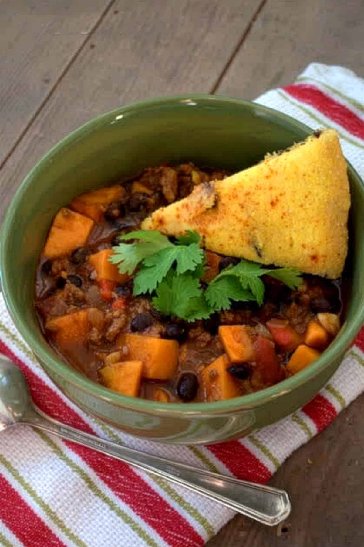 A green bowl of turkey chili with yams, black beans, sweet potatoes, and tomato pieces, garnished with fresh cilantro and a slice of cornbread. The bowl sits on a striped cloth napkin with a spoon beside it.