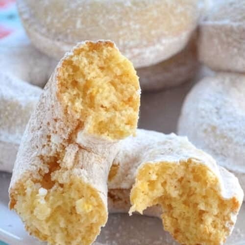 A close-up of baked donuts covered in powdered sugar on a plate, with one donut broken in half to reveal a soft, moist yellow inside. The plate rests on a colorful, floral-patterned tablecloth.