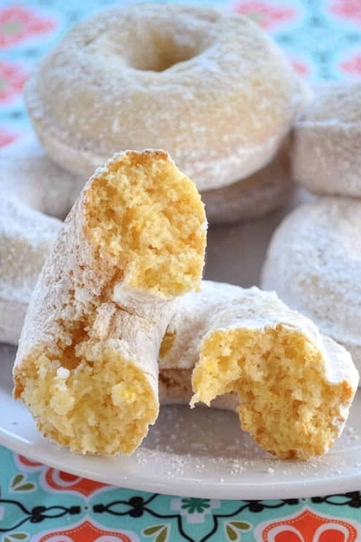 A close-up of baked donuts covered in powdered sugar on a plate, with one donut broken in half to reveal a soft, moist yellow inside. The plate rests on a colorful, floral-patterned tablecloth.