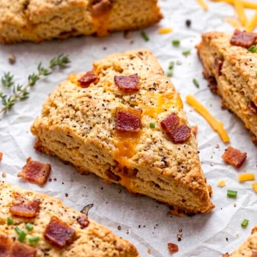 Golden brown, triangular Bacon Cheddar Scones topped with melted cheese, crispy bacon pieces, and green chives rest on parchment paper, surrounded by a sprig of thyme and scattered cheesy shreds.