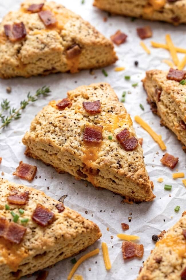 Golden brown, triangular Bacon Cheddar Scones topped with melted cheese, crispy bacon pieces, and green chives rest on parchment paper, surrounded by a sprig of thyme and scattered cheesy shreds.