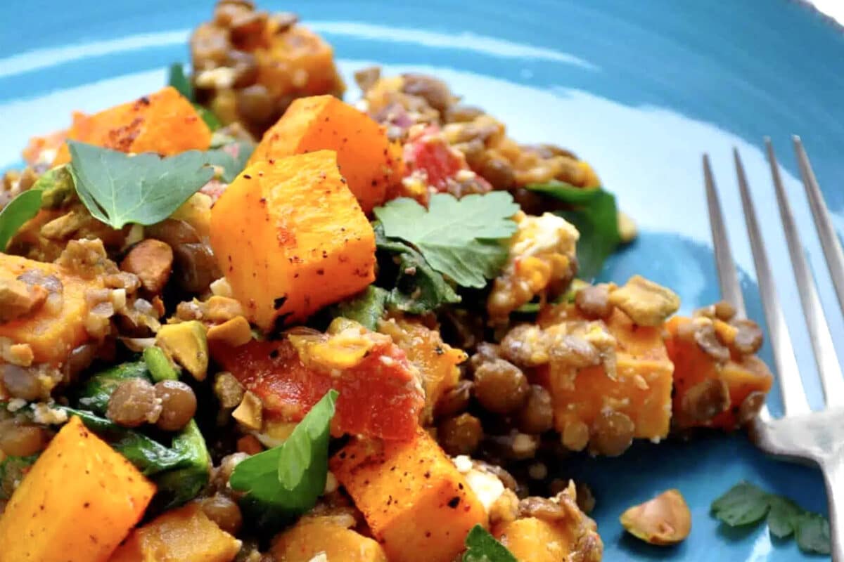 A close-up of a salad with warm butternut squash, lentils, red peppers, spinach, herbs, and nuts on a blue plate next to a fork.