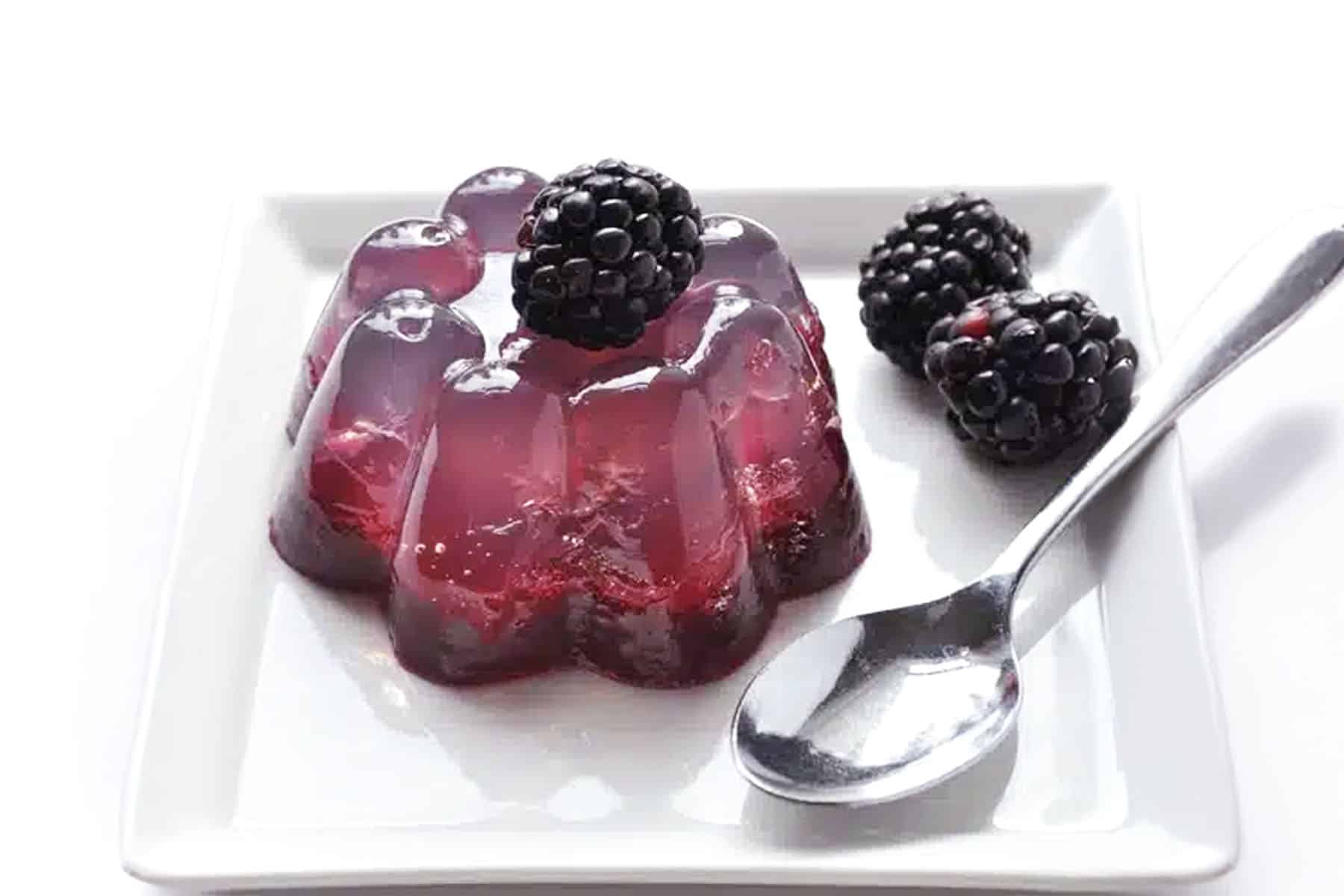 A plate with a flower-shaped purple coconut water gelatin dessert topped with a blackberry, two more blackberries on the side, and a metal spoon, all on a white square plate.