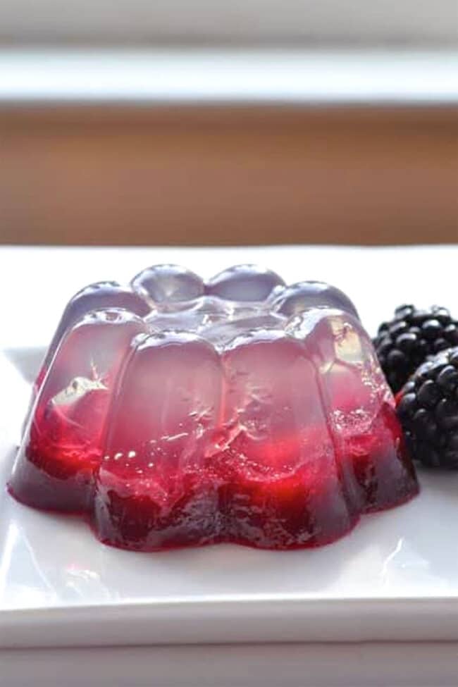 A clear and red coconut water gelatin dessert with berry pieces inside, molded into a flower shape, sits on a white plate next to two fresh blackberries.