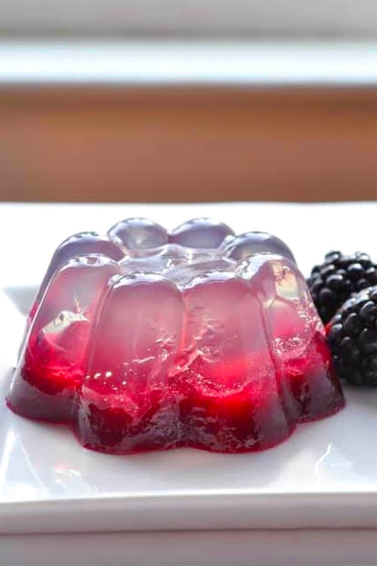 A clear and red coconut water gelatin dessert with berry pieces inside, molded into a flower shape, sits on a white plate next to two fresh blackberries.