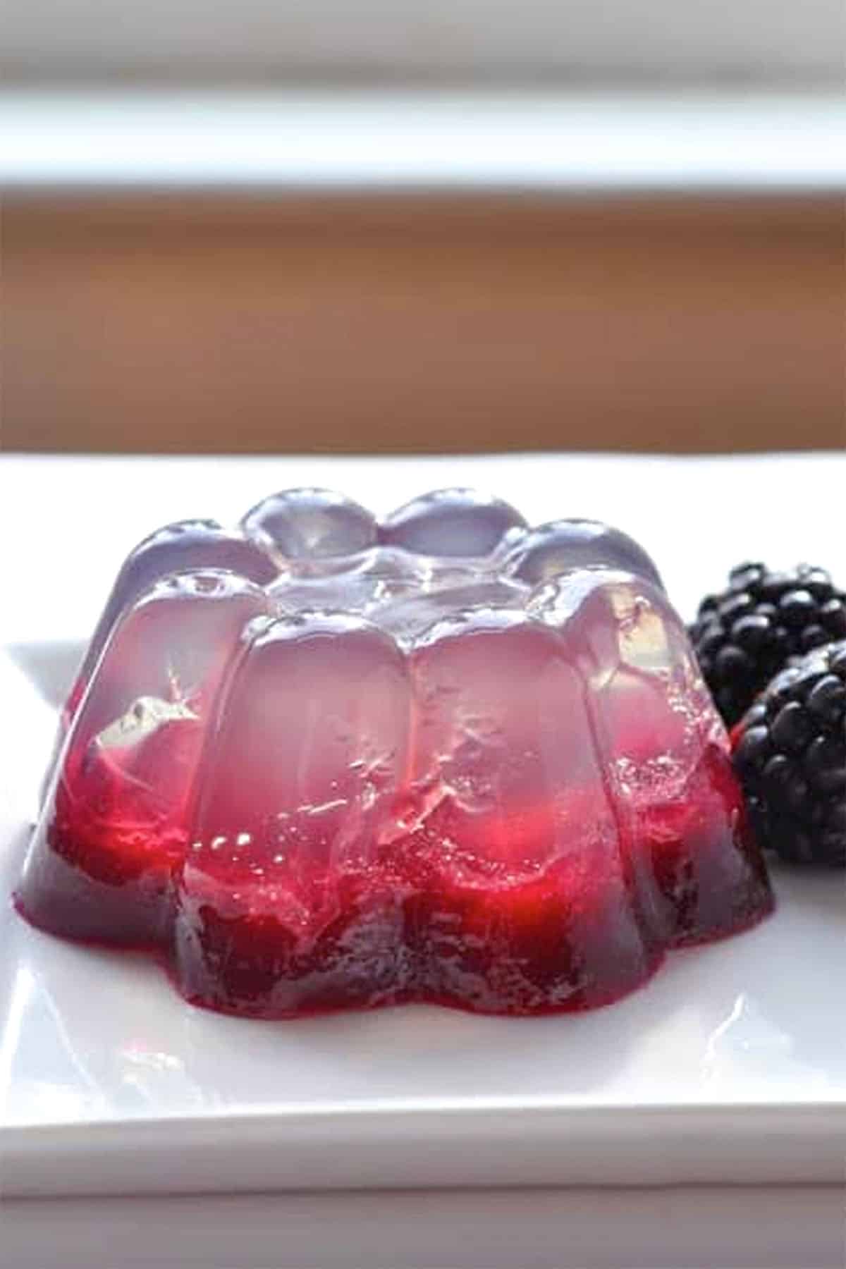 A clear and red coconut water gelatin dessert with berry pieces inside, molded into a flower shape, sits on a white plate next to two fresh blackberries.