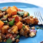A blue plate with a colorful lentil salad featuring warm butternut squash, spinach, herbs, and crumbled feta, with a fork on the side.