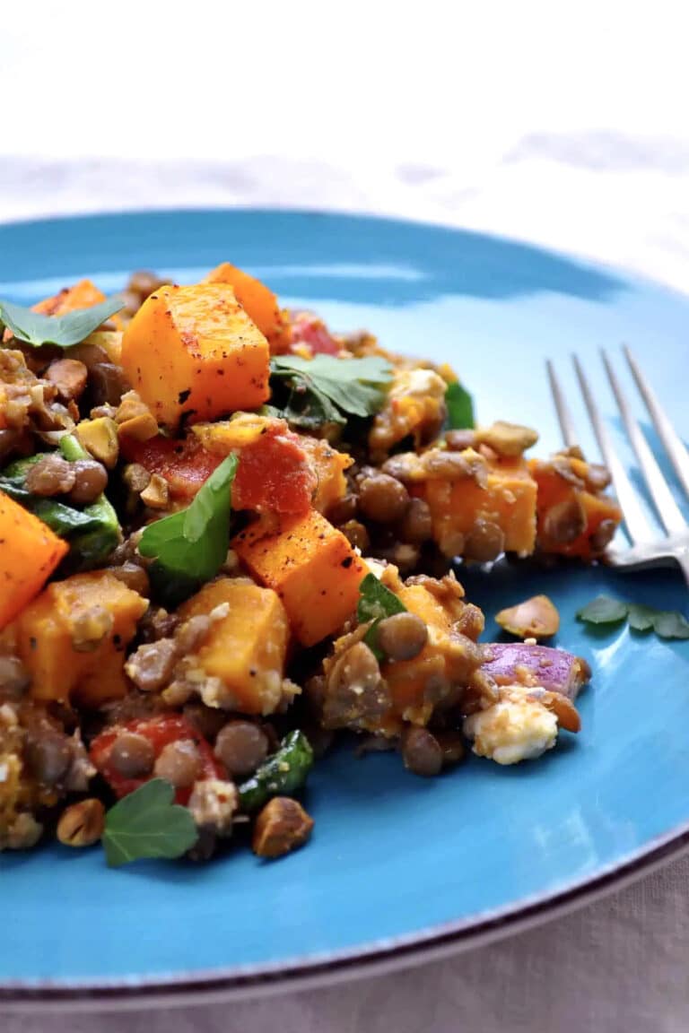 A blue plate with a colorful lentil salad featuring warm butternut squash, spinach, herbs, and crumbled feta, with a fork on the side.