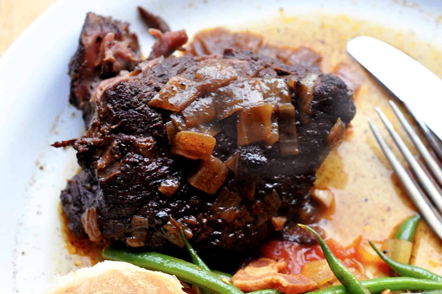 A plate with a serving of alcatra Portuguese pot roast topped with onions and sauce, accompanied by green beans, tomatoes, and a piece of bread on the side. A fork and knife rest on the plate.