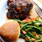 A plate with a piece of alcatra Portuguese pot roast topped with onions, sautéed green beans with bacon and tomatoes, and a round dinner roll, with a fork and knife to the side.