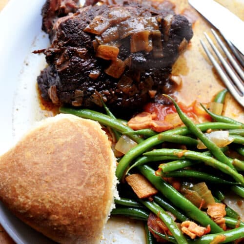 A plate with a piece of alcatra Portuguese pot roast topped with onions, sautéed green beans with bacon and tomatoes, and a round dinner roll, with a fork and knife to the side.