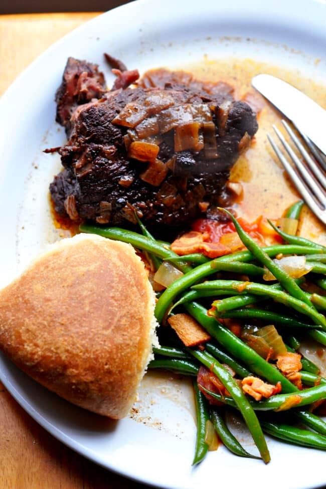 A plate with a piece of alcatra Portuguese pot roast topped with onions, sautéed green beans with bacon and tomatoes, and a round dinner roll, with a fork and knife to the side.