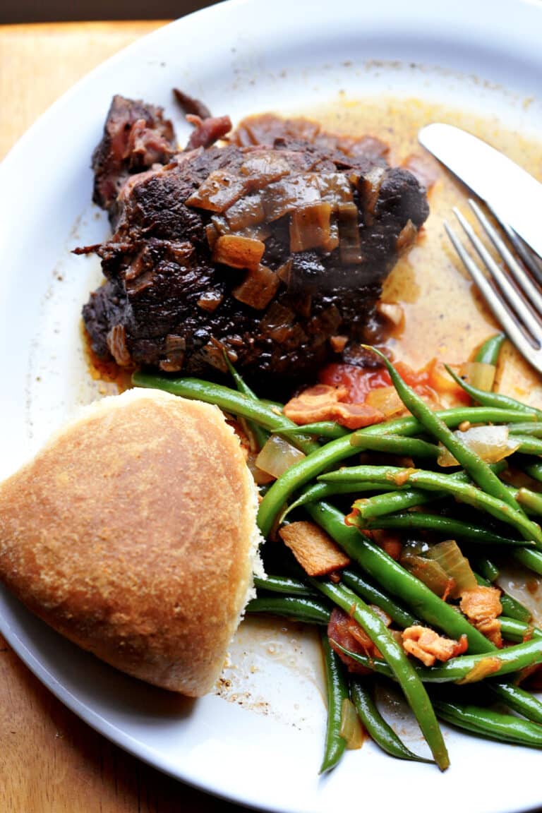 A plate with a piece of alcatra Portuguese pot roast topped with onions, sautéed green beans with bacon and tomatoes, and a round dinner roll, with a fork and knife to the side.