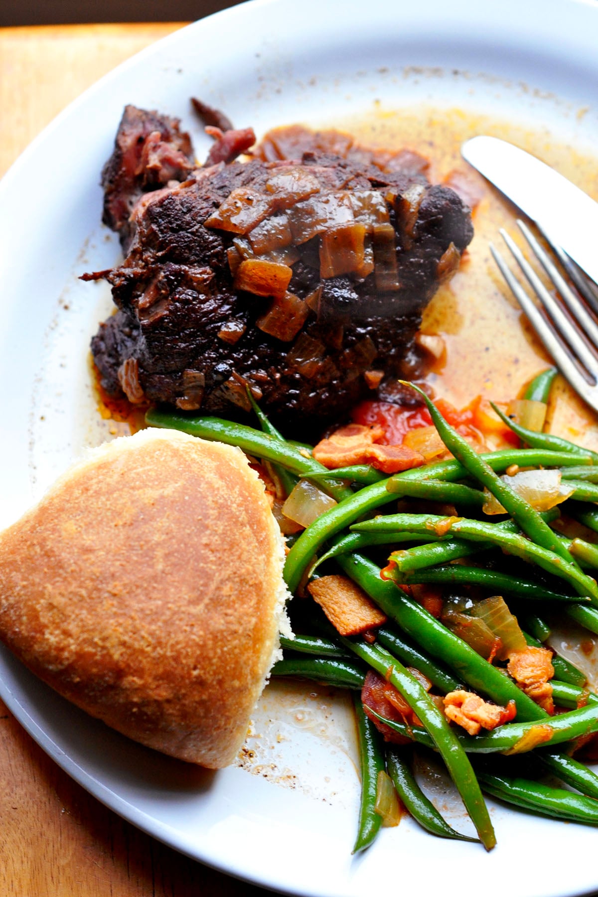 A plate with a piece of alcatra Portuguese pot roast topped with onions, sautéed green beans with bacon and tomatoes, and a round dinner roll, with a fork and knife to the side.