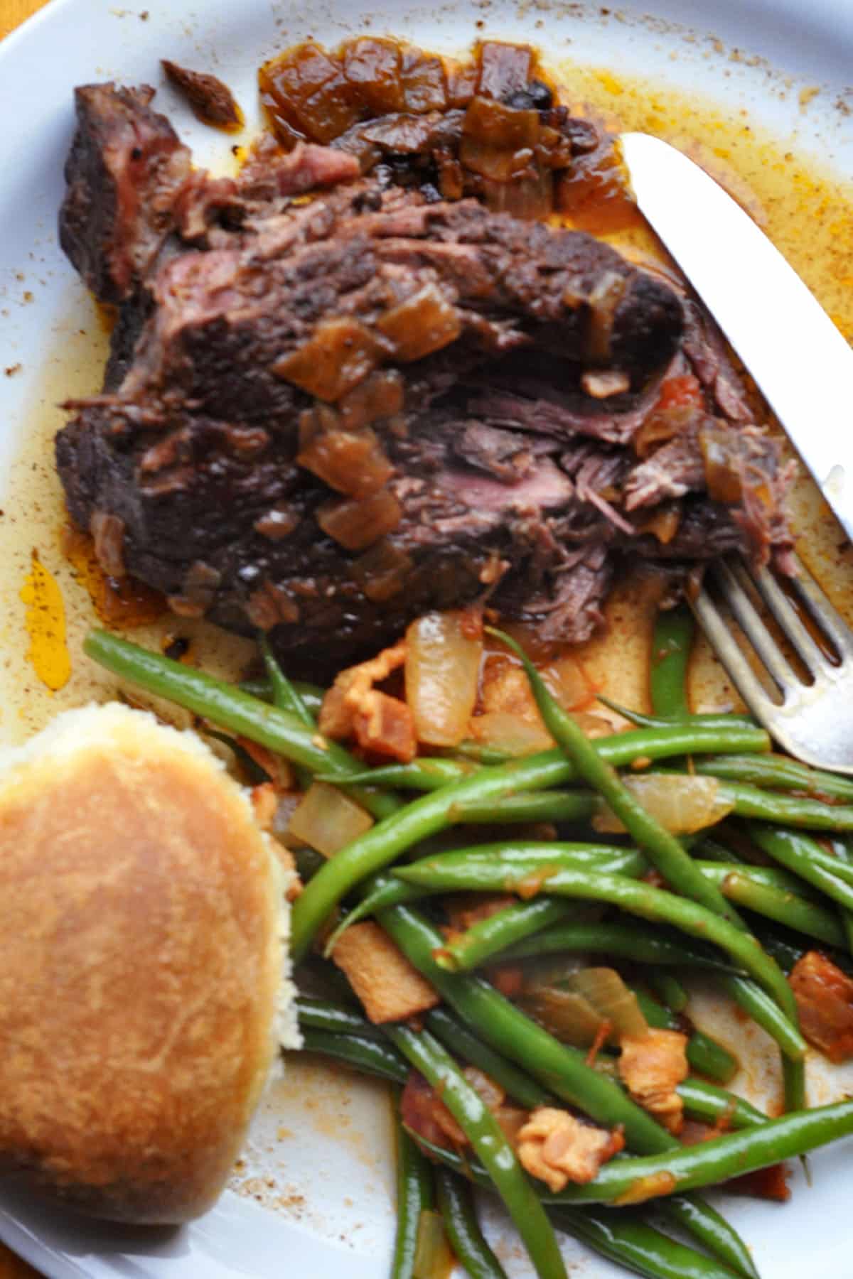 A plate with a serving of alcatra Portuguese pot roast topped with onions, green beans mixed with pieces of bacon, a dinner roll, and a fork and knife on the side.