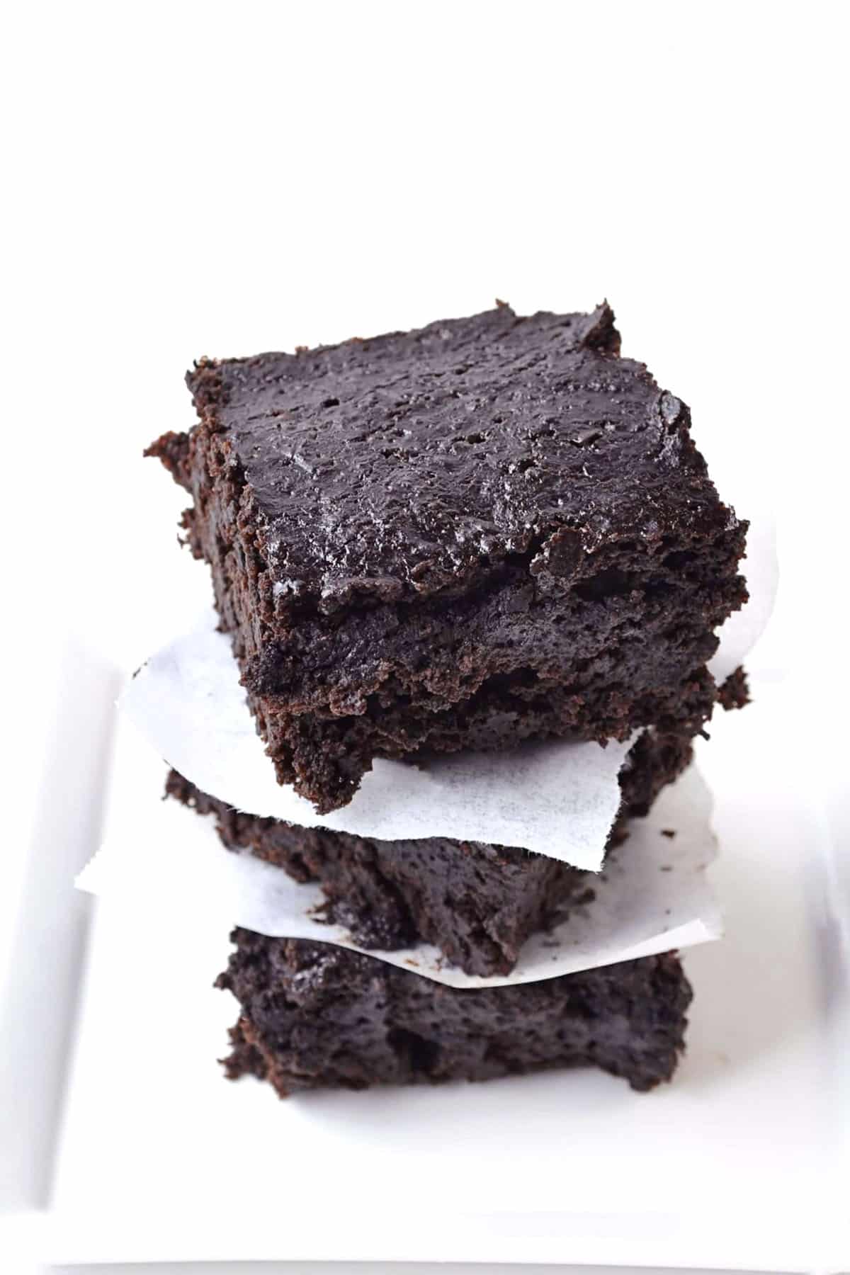 Three thick, fudgy chocolate stout brownies stacked on top of each other, separated by pieces of white parchment paper, on a white surface with a plain white background.
