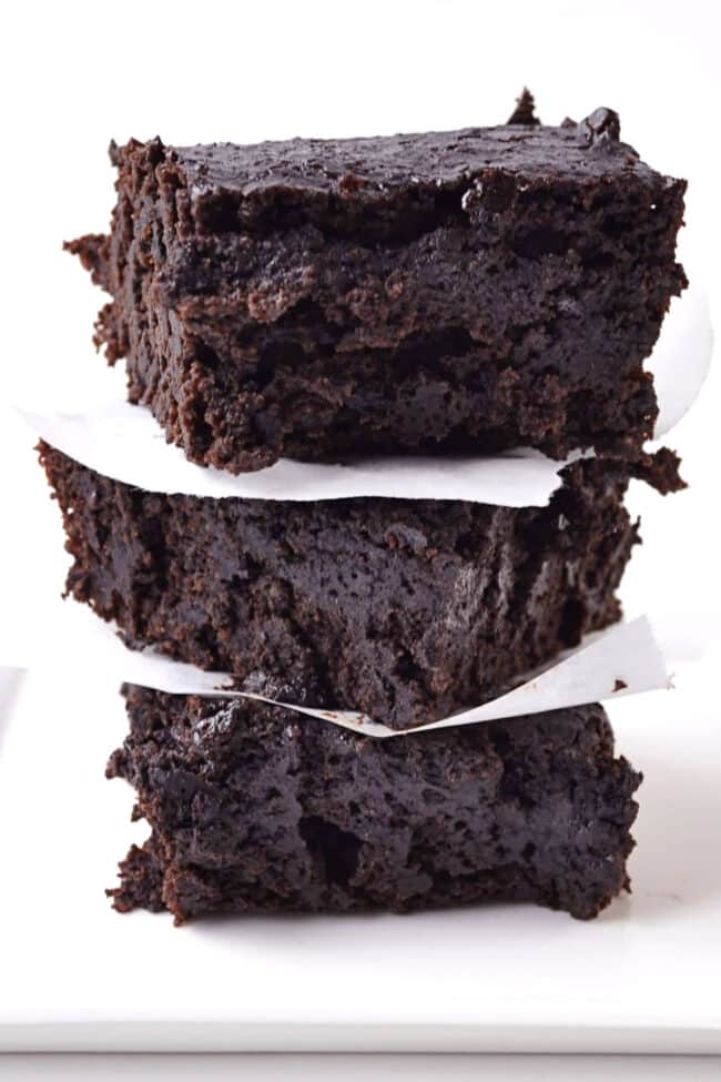 A stack of three thick, fudgy chocolate stout brownies separated by pieces of white parchment paper, set against a plain white background.