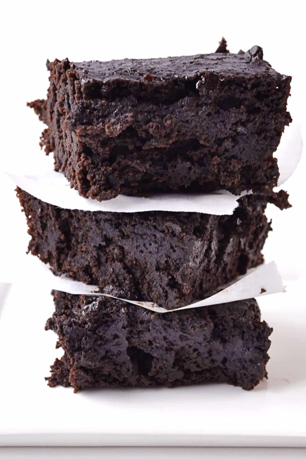 A stack of three thick, fudgy chocolate stout brownies separated by pieces of white parchment paper, set against a plain white background.