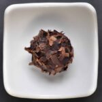 A single round chocolate truffle, crowned with delicate chocolate shavings, sits in the center of a small, square white dish on a dark background—an elegant showcase of artisanal truffles.