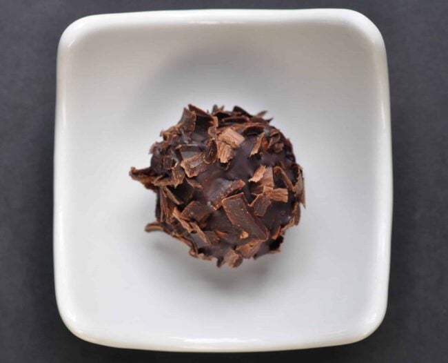 A single round chocolate truffle, crowned with delicate chocolate shavings, sits in the center of a small, square white dish on a dark background—an elegant showcase of artisanal truffles.