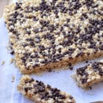 A batch of chocolate chip granola bars topped with mini chocolate chips sits on parchment paper. One bar has been cut and separated from the rest, with a knife placed nearby on a wooden surface.