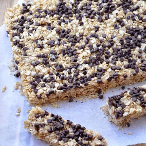 A batch of chocolate chip granola bars topped with mini chocolate chips sits on parchment paper. One bar has been cut and separated from the rest, with a knife placed nearby on a wooden surface.