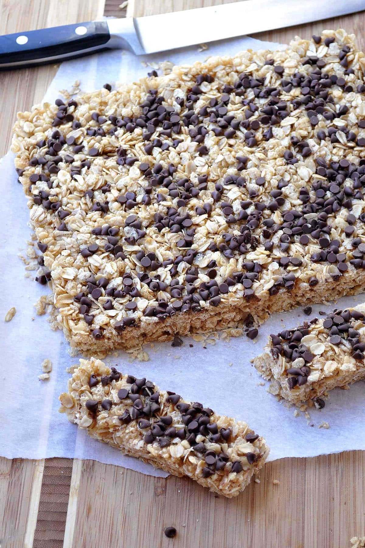A batch of chocolate chip granola bars topped with mini chocolate chips sits on parchment paper. One bar has been cut and separated from the rest, with a knife placed nearby on a wooden surface.