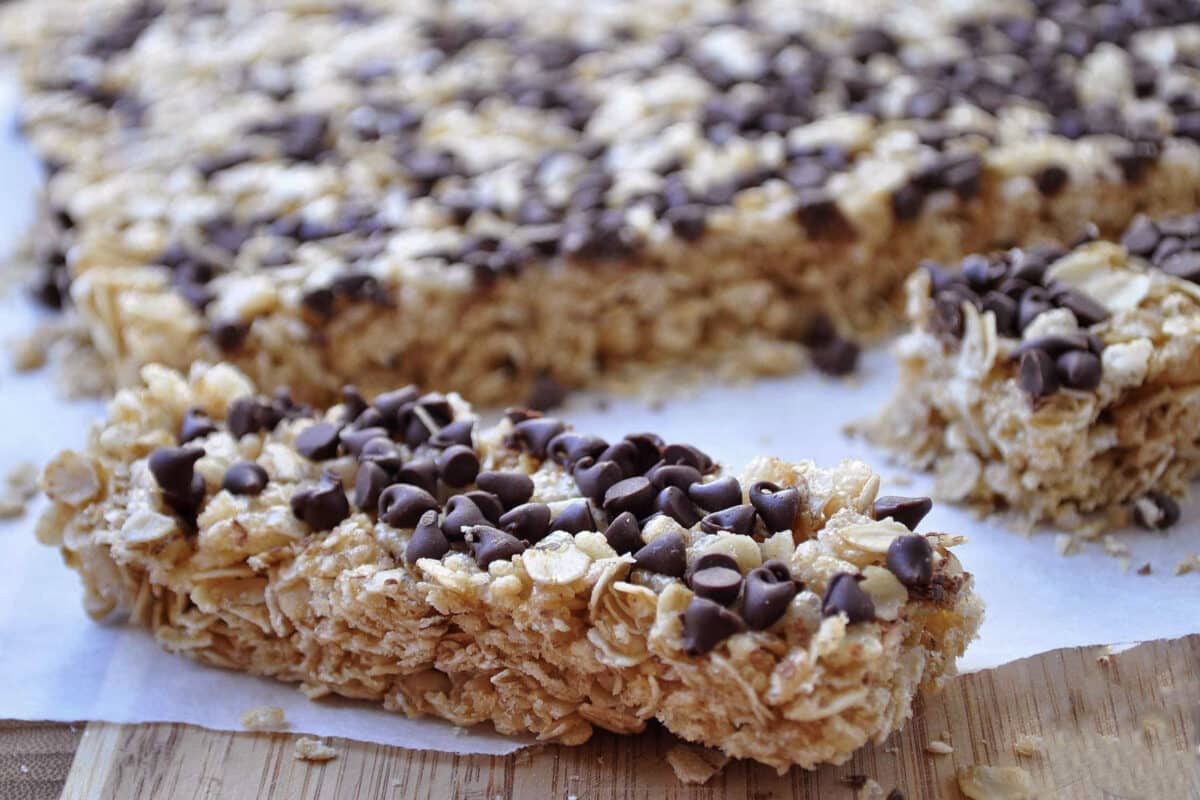 A close-up of homemade chocolate chip granola bars topped with extra chips, with one bar cut and placed in the foreground on a wooden surface. The chewy bars rest on parchment paper, showcasing their delicious texture.