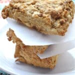 Three golden-brown cinnamon yogurt scones are stacked on a white plate, separated by pieces of parchment paper, with a colorful floral tablecloth underneath.