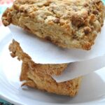 Two thick, golden-brown cinnamon yogurt scones are stacked on a white plate, separated by sheets of parchment paper. The plate sits on a colorful patterned tablecloth near a bright window.