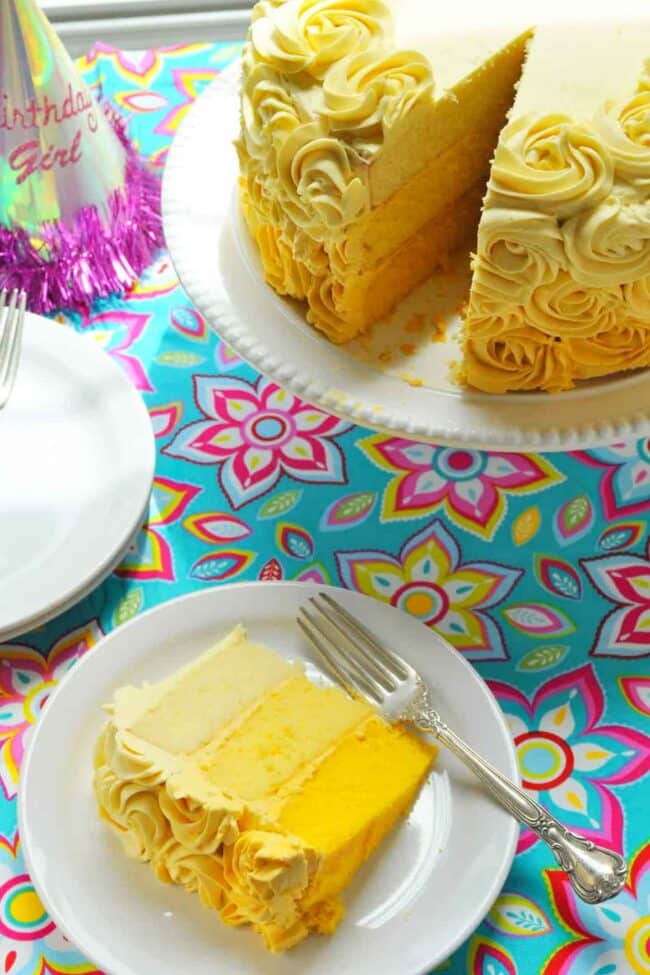 A slice of vanilla cake with creamy frosting sits on a white plate with a fork. The cake, decorated with frosting rosettes, is on a white stand. A colorful tablecloth and a “Birthday Girl” party hat are also visible.