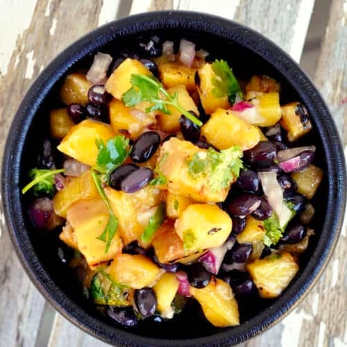 A black bowl filled with a vibrant grilled pineapple salsa—pineapple chunks, black beans, red onion, and cilantro—rests on a rustic white wooden surface.