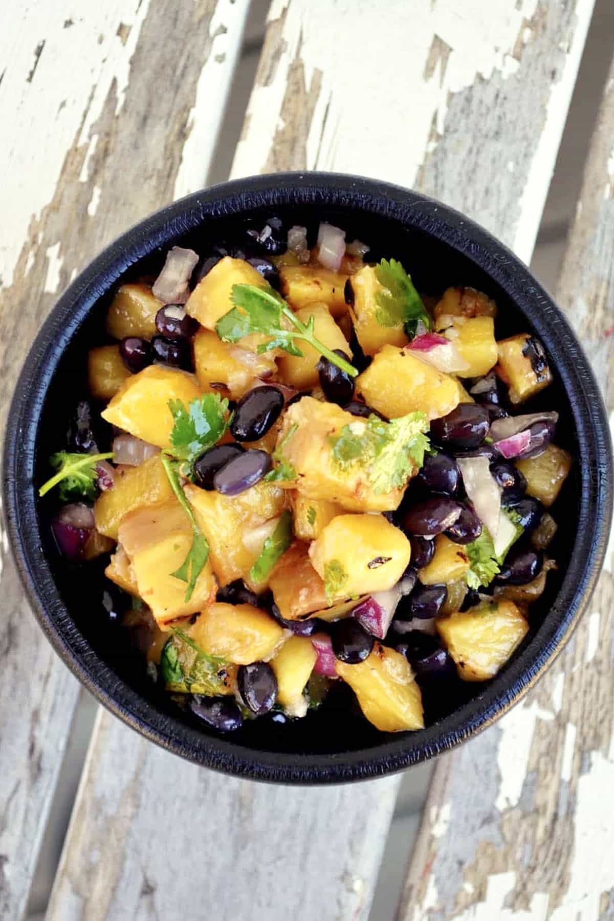 A black bowl filled with a vibrant grilled pineapple salsa—pineapple chunks, black beans, red onion, and cilantro—rests on a rustic white wooden surface.
