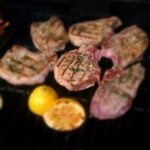 Grilled pork chops with charred grill marks and herbs sizzle on a barbecue, paired perfectly with halved lemons—an ideal match for a fresh apple parmesan salad.