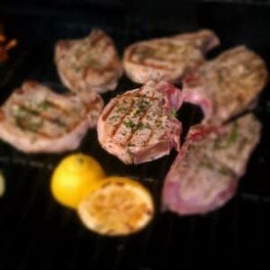 Grilled pork chops with charred grill marks and herbs sizzle on a barbecue, paired perfectly with halved lemons—an ideal match for a fresh apple parmesan salad.