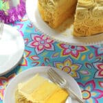 A slice of yellow layered vanilla cake with yellow and white rosette frosting sits on a white plate with a fork. The rest of the cake is on a stand nearby, and a Birthday Girl party hat is visible on a colorful floral tablecloth.