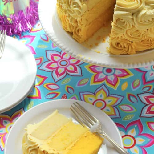 A slice of yellow layered vanilla cake with yellow and white rosette frosting sits on a white plate with a fork. The rest of the cake is on a stand nearby, and a Birthday Girl party hat is visible on a colorful floral tablecloth.