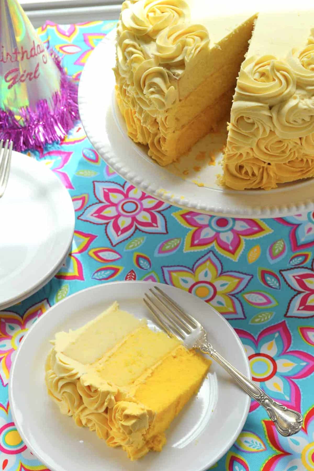A slice of yellow layered vanilla cake with yellow and white rosette frosting sits on a white plate with a fork. The rest of the cake is on a stand nearby, and a Birthday Girl party hat is visible on a colorful floral tablecloth.