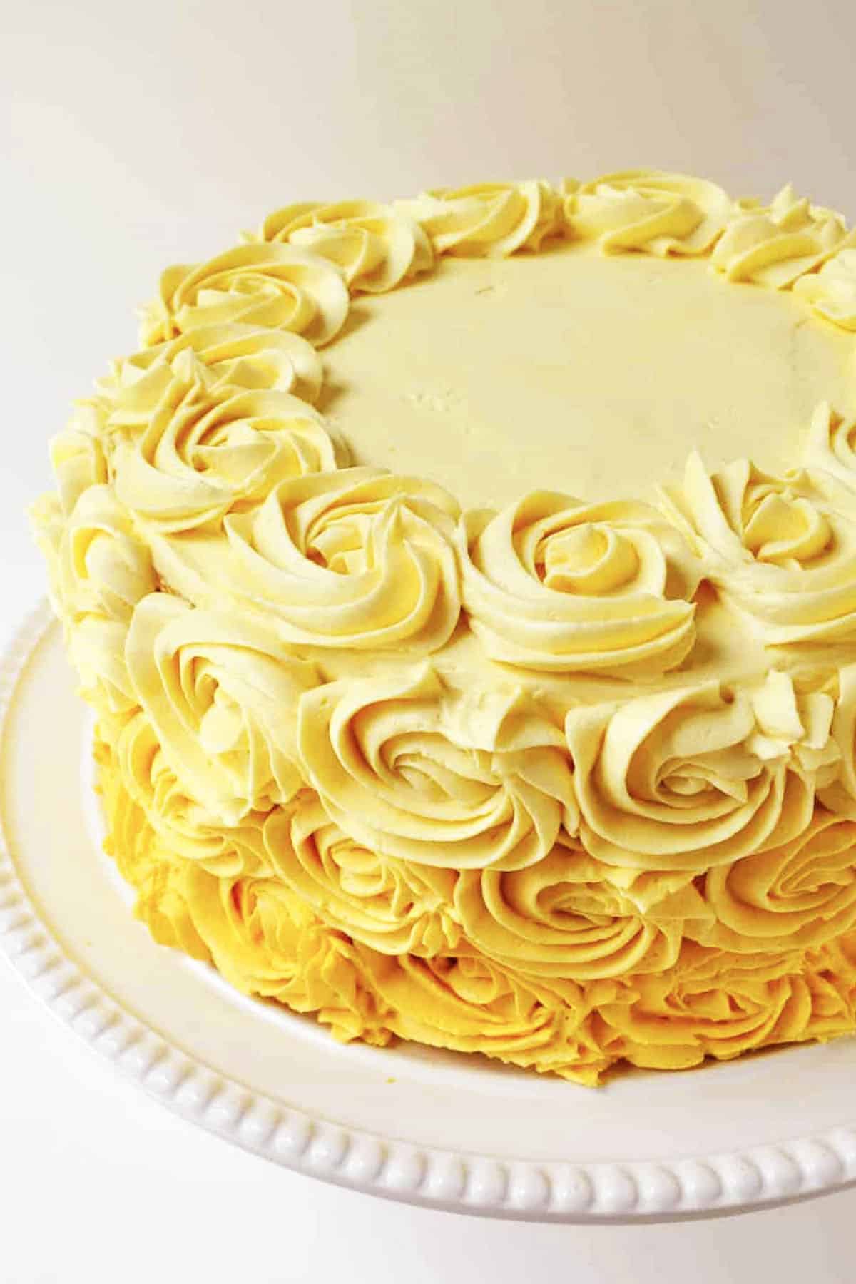 A round vanilla cake decorated with yellow buttercream rosettes on the sides and top edge, displayed on a white plate. The center of the cake’s top is smooth and unfrosted.