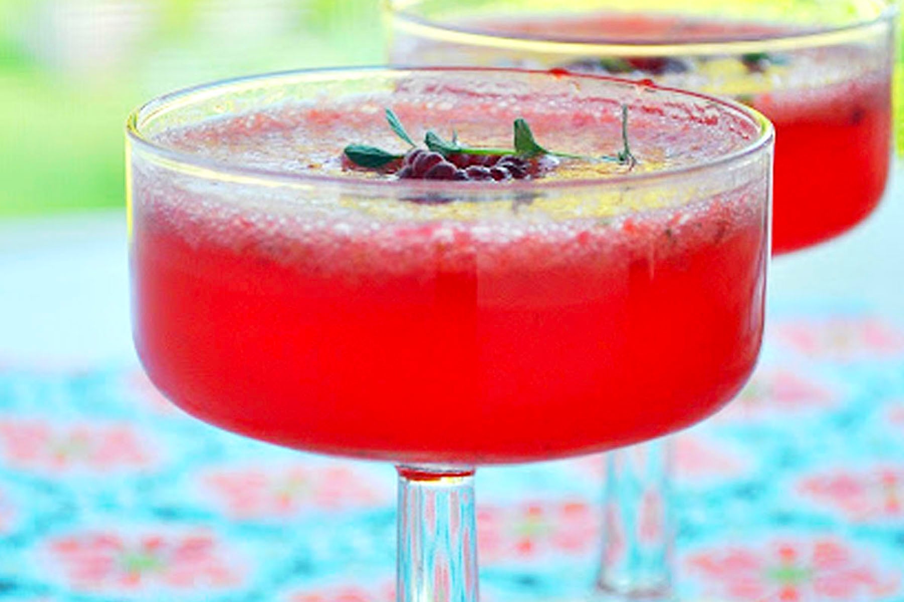 A close-up of two coupe glasses filled with a bright red, frothy berry gin cocktail, garnished with berries and fresh herbs, set on a colorful patterned surface.