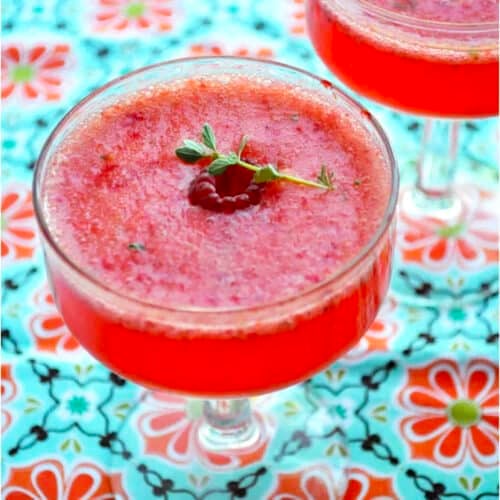 Two glass cups filled with a vibrant berry Tom Collins, garnished with fresh raspberries and green herbs, sit on a colorful floral-patterned tablecloth.