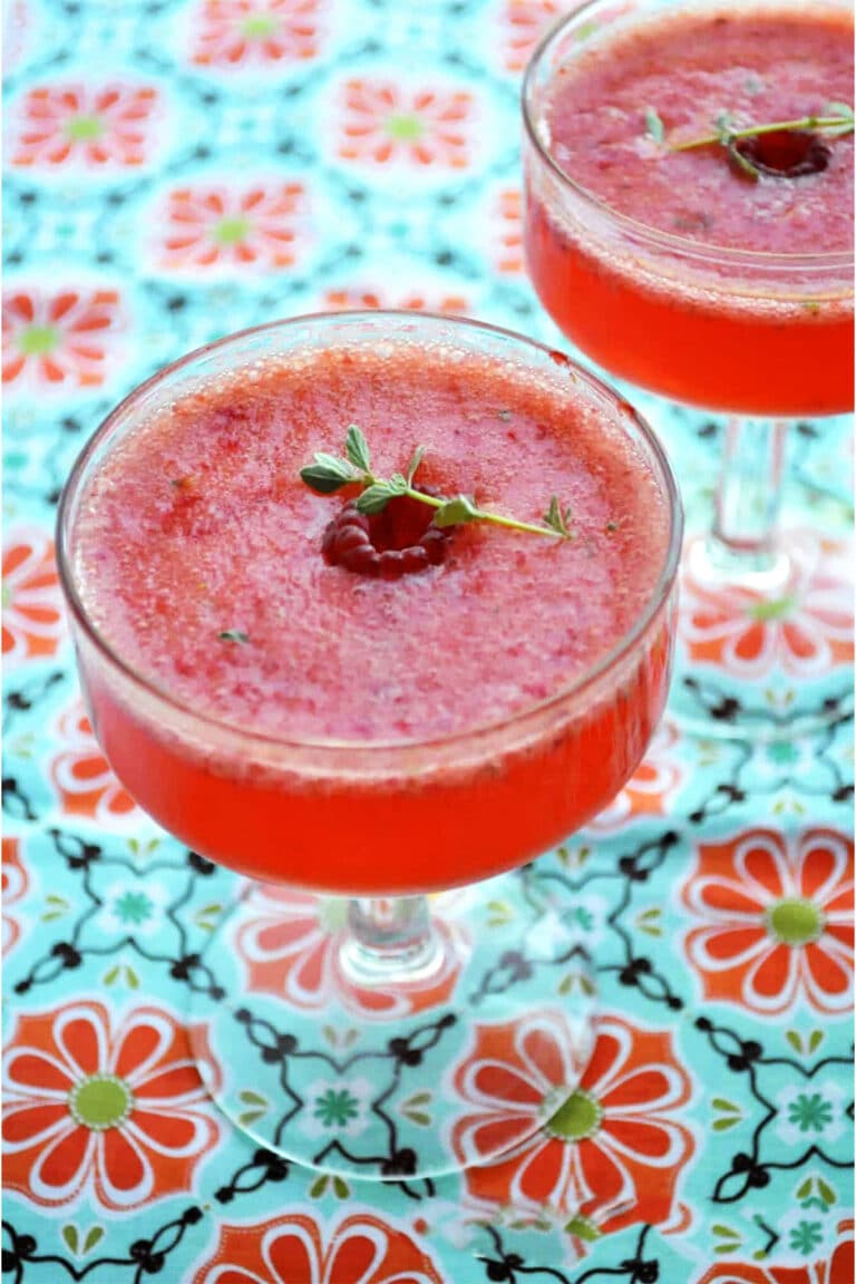 Two glass cups filled with a vibrant berry Tom Collins, garnished with fresh raspberries and green herbs, sit on a colorful floral-patterned tablecloth.