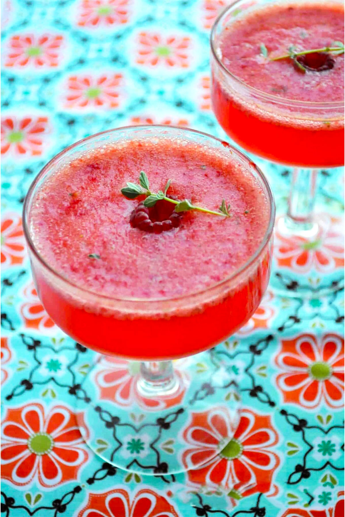 Two glass cups filled with a vibrant berry Tom Collins, garnished with fresh raspberries and green herbs, sit on a colorful floral-patterned tablecloth.