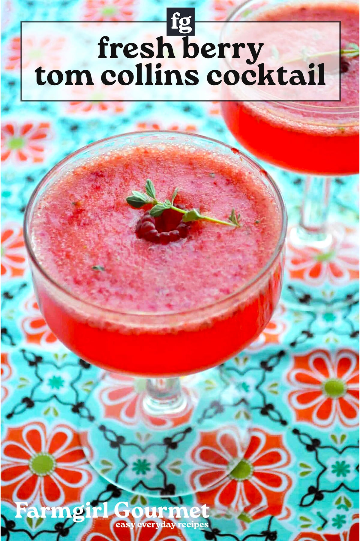 Two glasses of bright pink berry Tom Collins cocktails, garnished with a raspberry and herb sprig, sit on a colorful floral-patterned tablecloth. Text overlay describes the berry Tom Collins drink and its source, Farmgirl Gourmet.