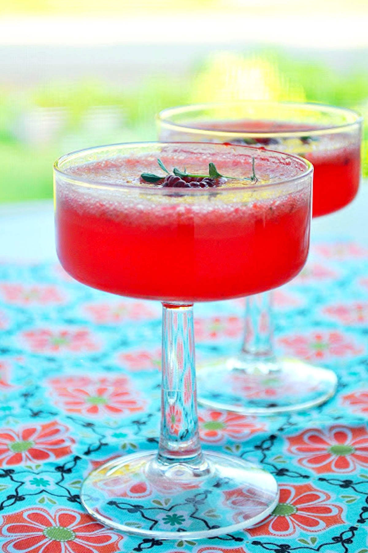 Two coupe glasses filled with a bright red, frothy berry Tom Collins, garnished with small green herbs, sit on a colorful floral-patterned tablecloth with a blurred outdoor background.