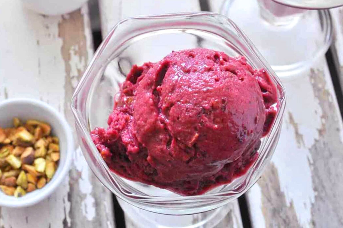 A close-up of a glass dish holding a scoop of deep purple Quick Cherry Ice Cream, with a small bowl of chopped pistachios nearby on a rustic white wooden surface.