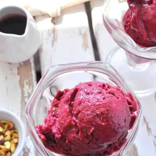 Two glass dishes filled with scoops of deep purple berry sorbet and Quick Cherry Ice Cream sit on a white wooden surface. A small bowl of pistachios and a pitcher of dark syrup nearby glisten in the bright natural light.
